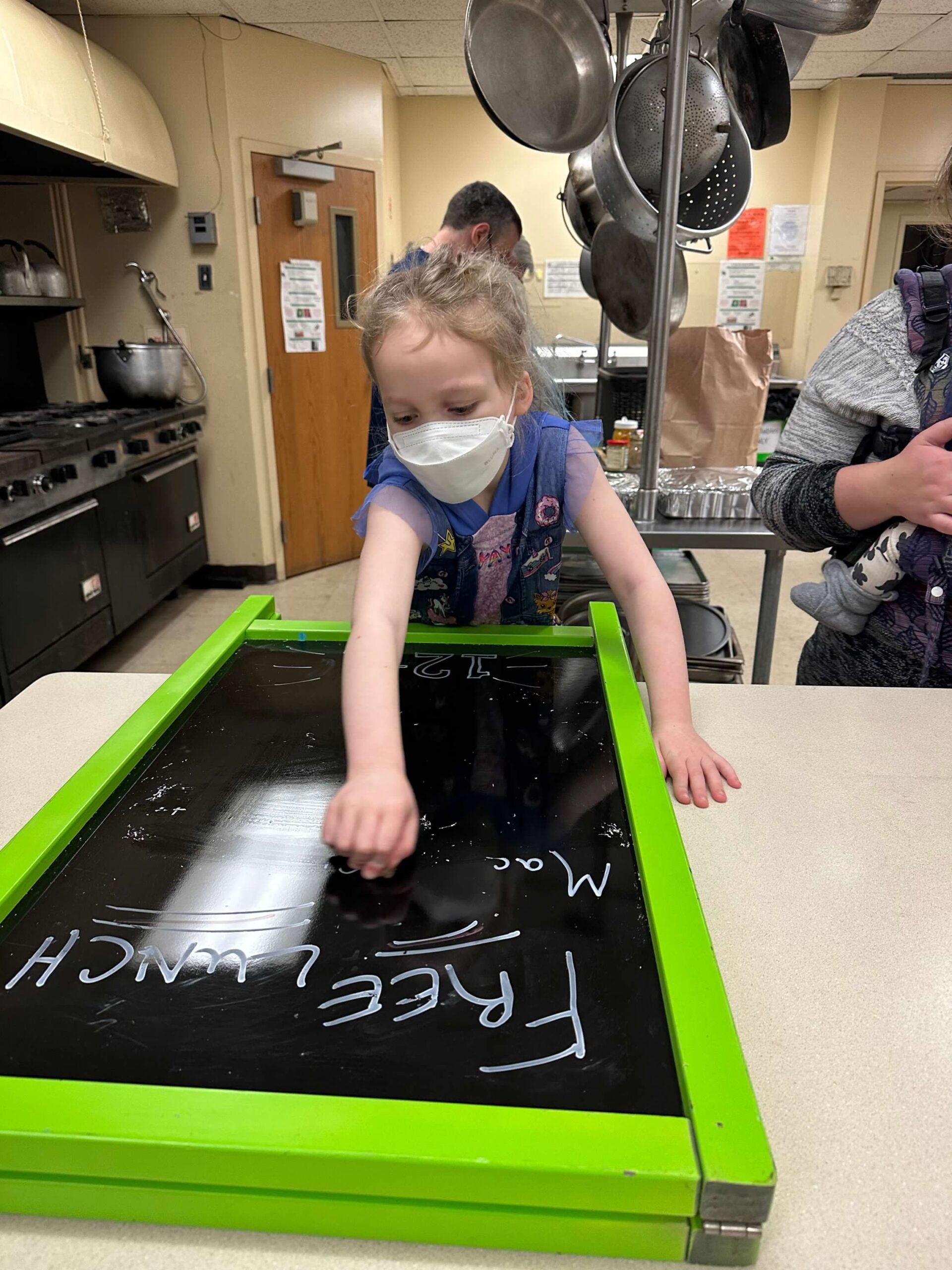 A small child wearing a mask cleans a sandwich board that says free lunch.