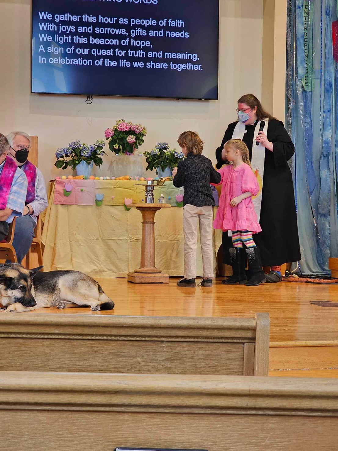 Two children help Reverend Molly light the chalice. A table with a pastel tablecloth and flowers is behind them.