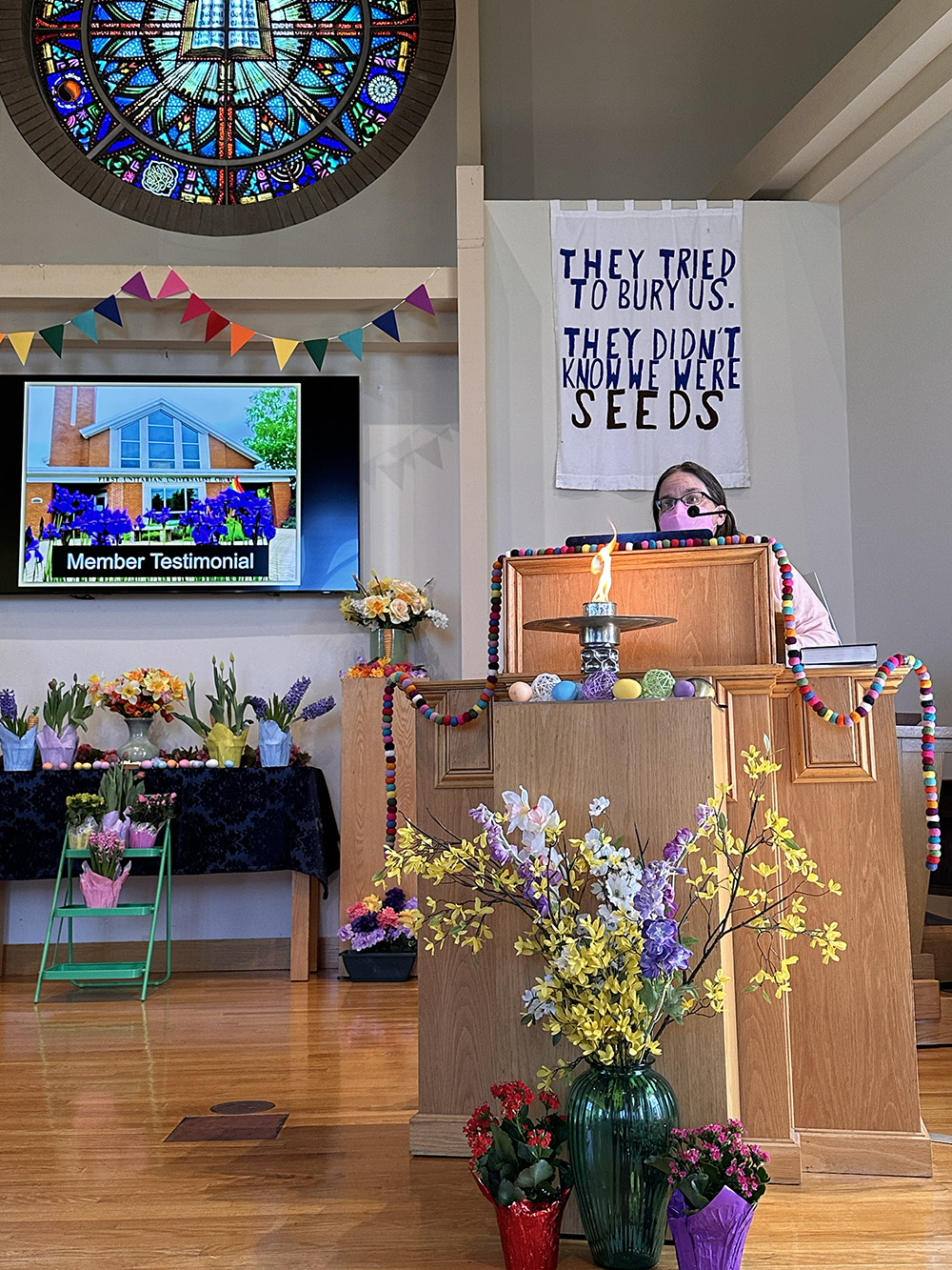 A person stands at the podium, wearing a pink mask. There is a large sign behind the podium that says “They tried to bury us. They didn’t know we were seeds.” A chalice is lit in front of the podium. There are colorful flowers all around the chancel. Rainbow colored flags hang above a large screen that says “member testimonial“.