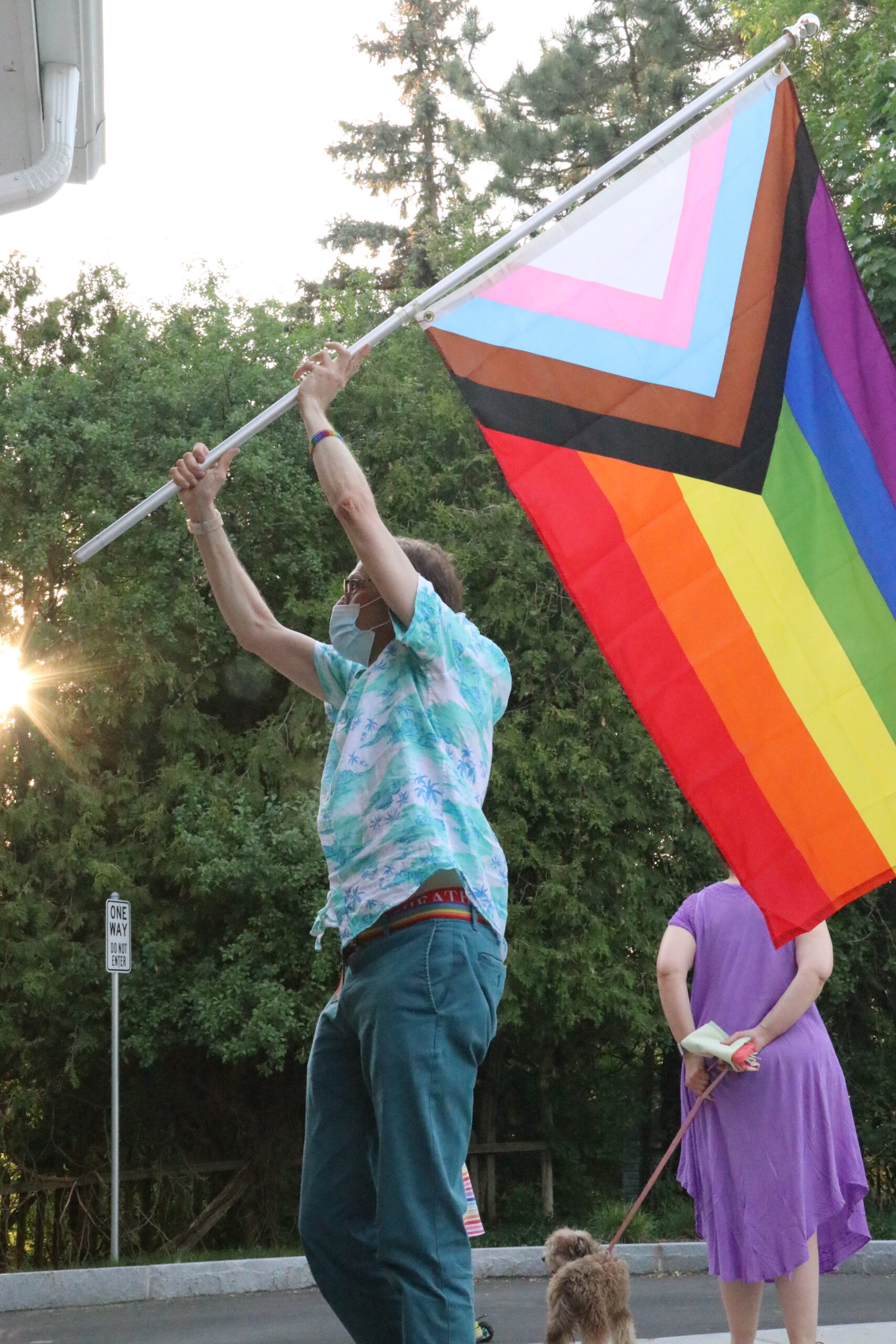 A masked person wearing a green and white shirt and green pants holds a Progress Pride flag on a pole in their hands above their head.