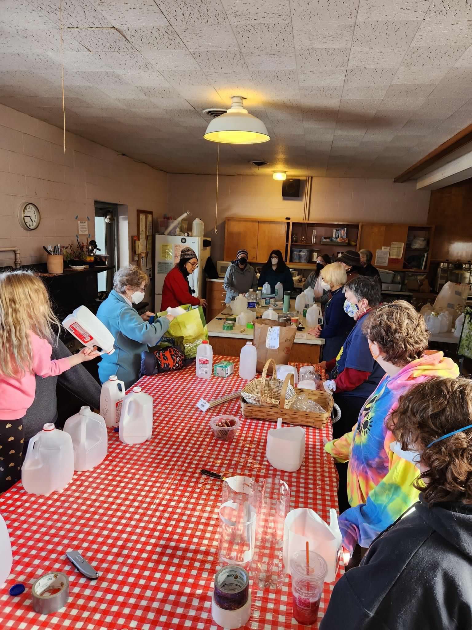 Many masked people of all ages gather around large tables in a kitchen. On the tables are empty gallon jugs, dirt and seeds.
