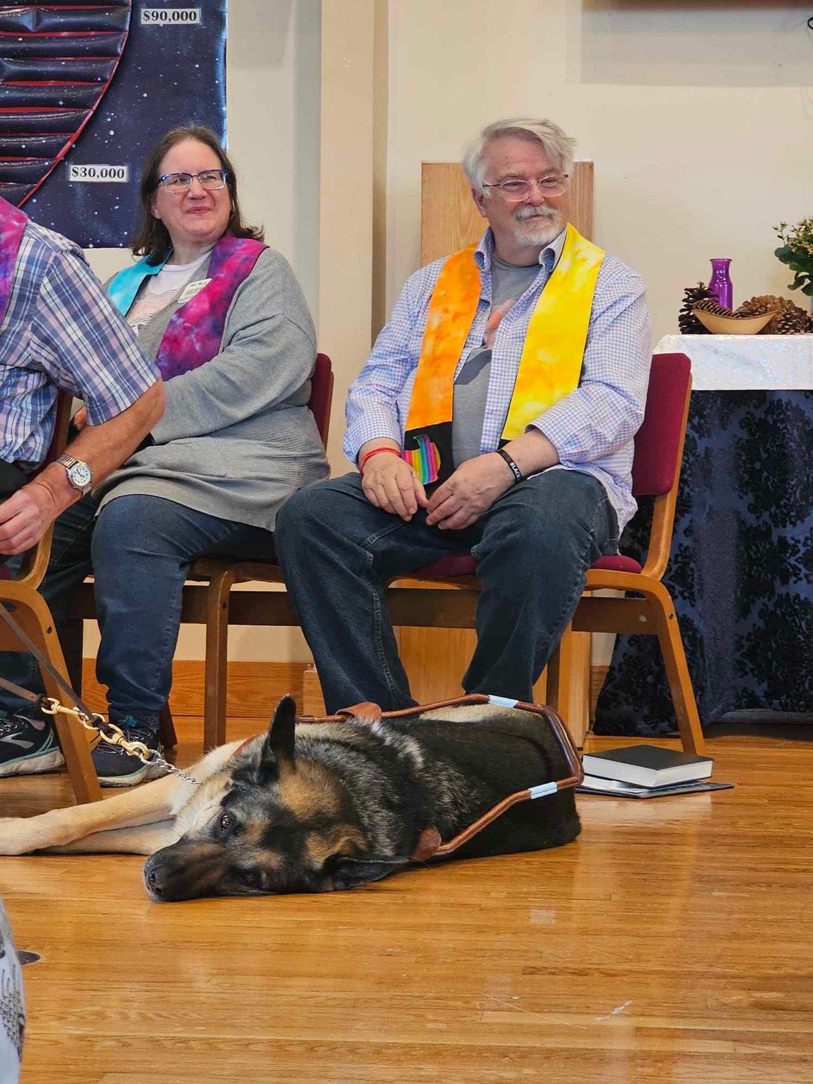 Two people with light skin who are wearing colorful stoles smile. At their feet a brown and black seeing eye dog lies on his side resting.