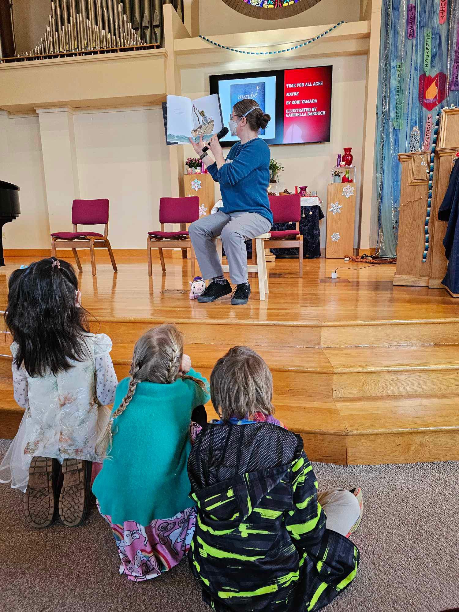 A masked person sits on a stool on the chancel holding a picture book and a microphone. Several children sit on the steps in the front listening.