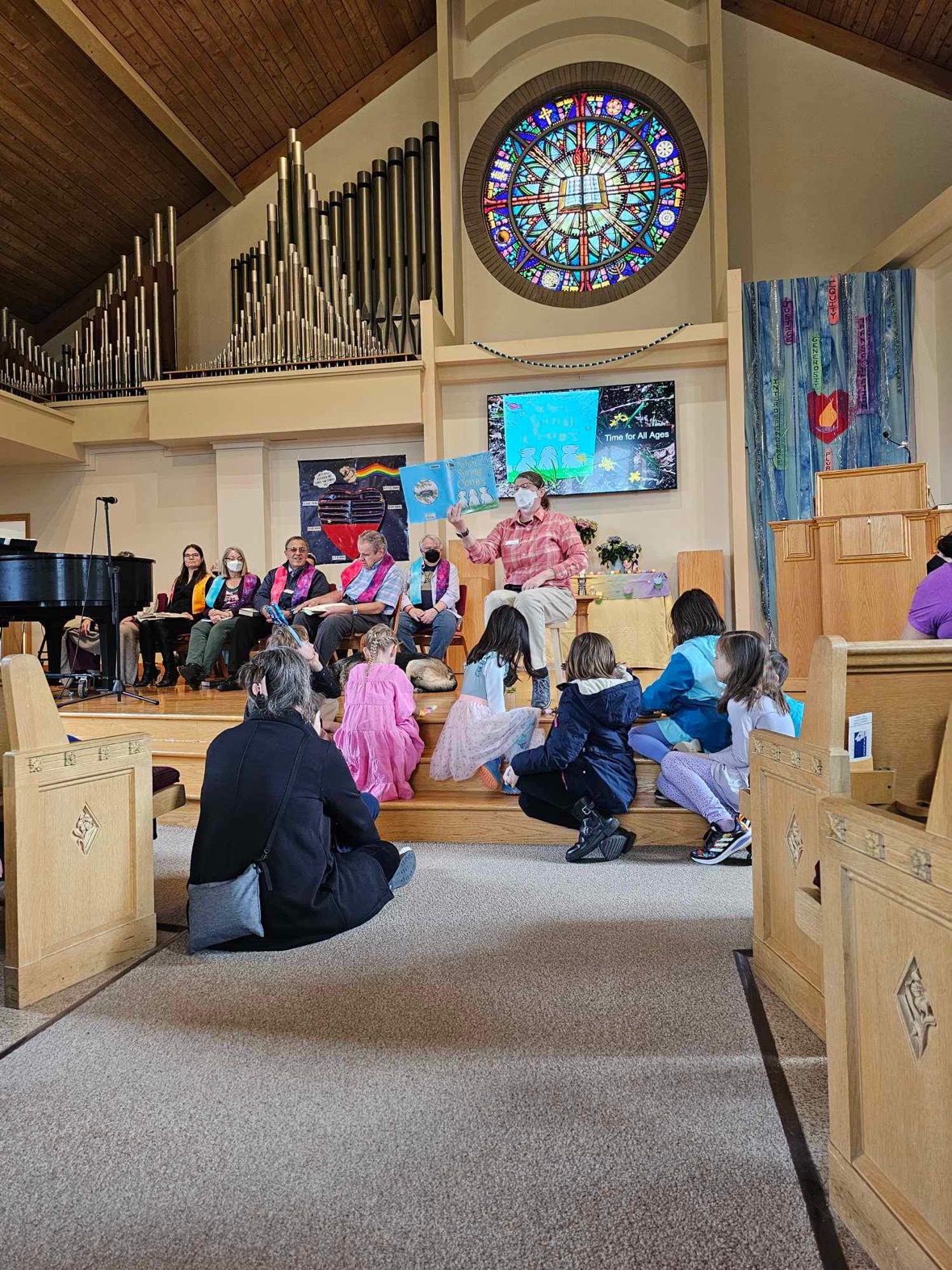 A masked person sits on a stool on the chancel holding a picture book and a microphone. Several children and adults sit on the steps in the front listening. The choir is seated behind.