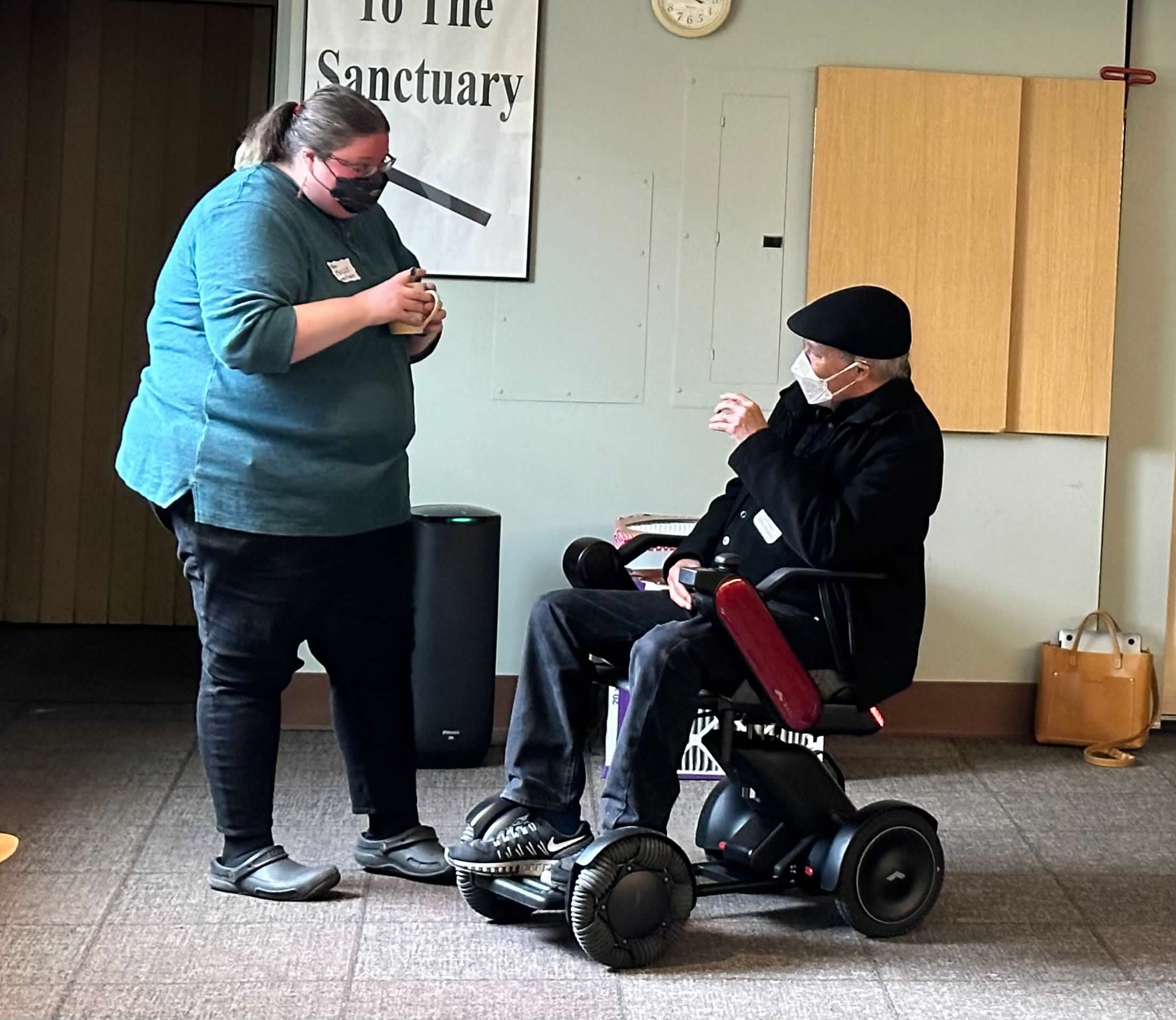 Two masked people have a conversation. One is standing and the other is sitting in a motorized wheelchair