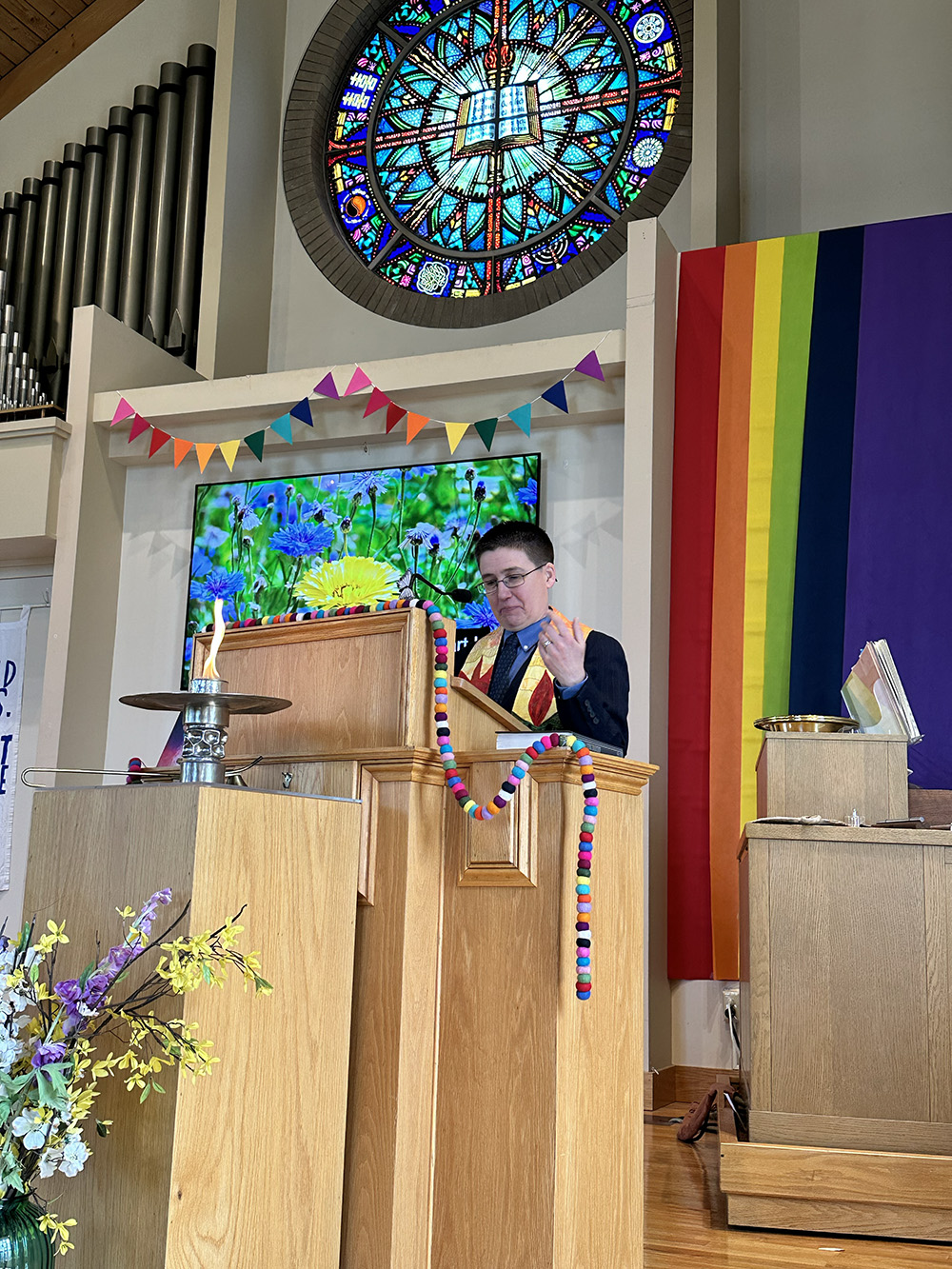 A light skinned person with short brown hair stands at the pulpit wearing a colorful stole. A large rainbow is behind them and a lit chalice is in front of them.