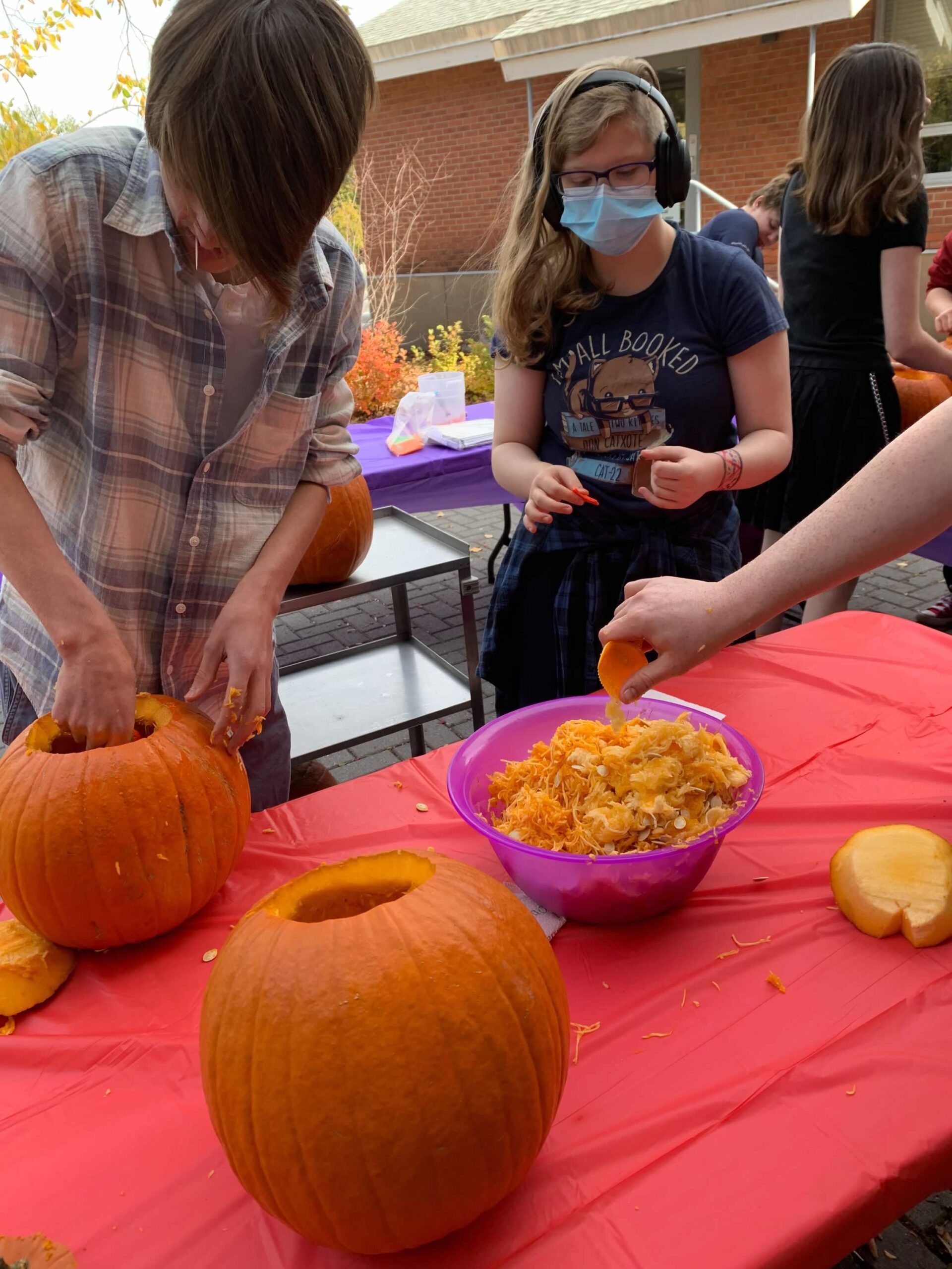 A group of teenagers gather around a red table carving pumpkins. There is a bowl in the middle of the table with seeds and pulp that has been scooped out of the pumpkins.