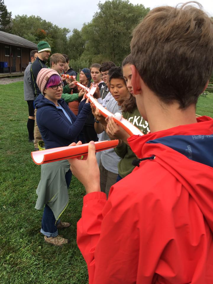 Teens and adults stand in a field in a line while holding pieces of gutter. They are working together to move a small ball from one end of the gutter to the other.