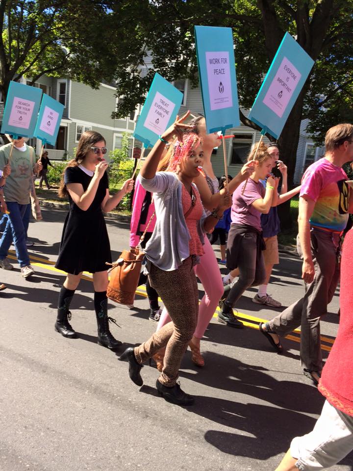 A group of teenagers walk in a parade carrying signs and playing kazoos.