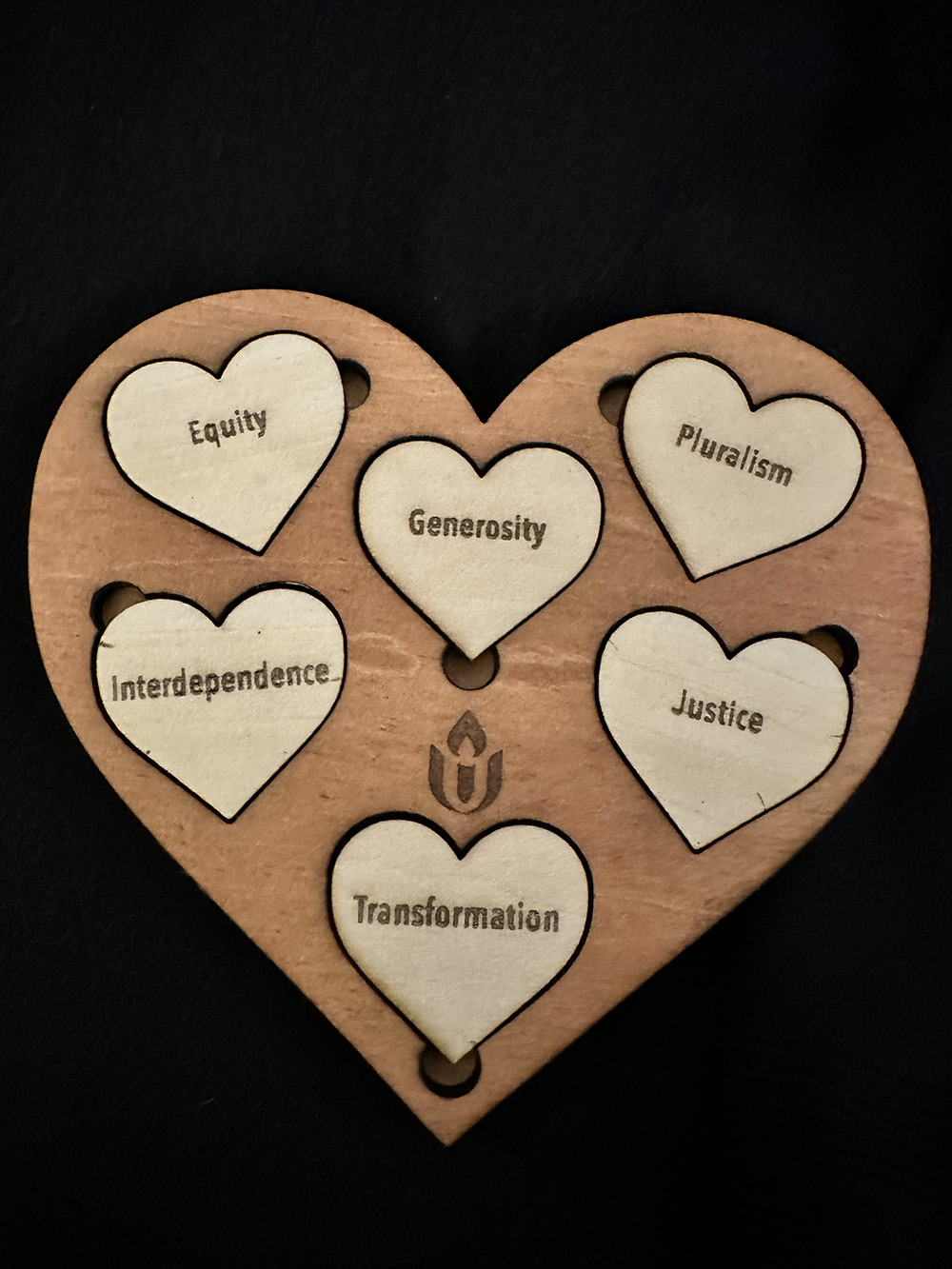 Wooden Heart-shaped puzzle with six smaller heart-shaped pieces. Each piece has one word. Equity, generosity, pluralism, interdependence, justice, transformation.