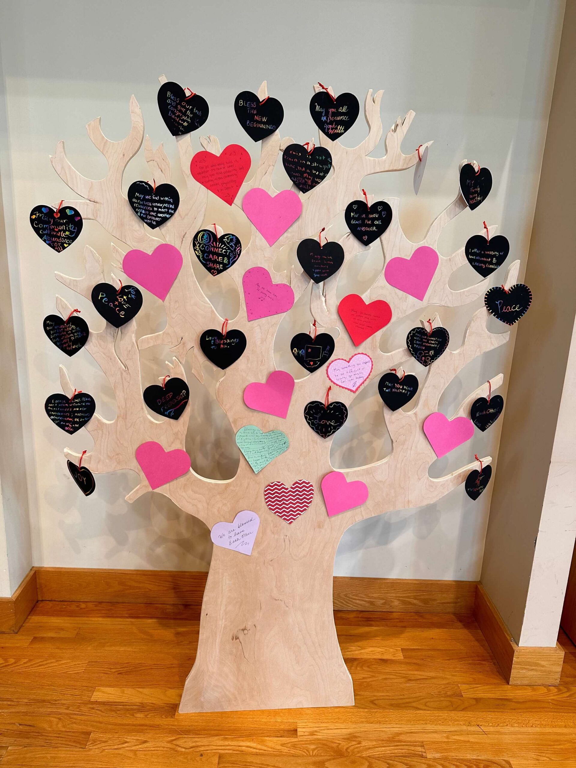 A large wooden tree cut out covered with black red, pink and green paper hearts.